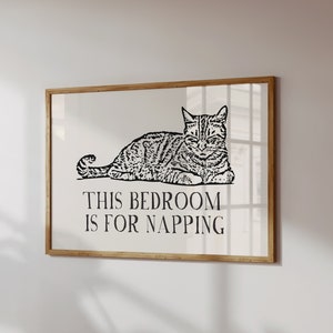 May include: A black and white illustration of a cat napping with the text "This bedroom is for napping" printed below. The illustration is framed in a light wood frame.