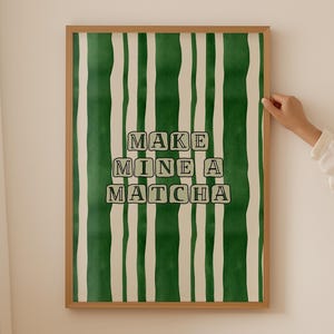 May include: A rectangular wall art print in a light wood frame featuring a vertical pattern of alternating dark green and cream-coloured wavy stripes. The centre displays the text "MAKE MINE A MATCHA" in black capital letters inside cream boxes.