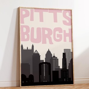 Könnte beinhalten: Gerahmter Kunstdruck mit einer stilisierten Silhouette der Skyline von Pittsburgh in Grau- und Schwarztönen. Das Wort "PITTSBURGH" ist in großen, rosa Buchstaben auf cremefarbenem Hintergrund geschrieben. Der Druck ist in einem Holzrahmen.