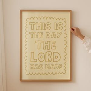 May include: A rectangular wall art print in a light wood frame featuring a cream-colored background with a scalloped border and the brown text THIS IS THE DAY THE LORD HAS MADE written in a bold, hand-drawn, rounded sans-serif typeface.