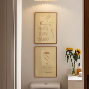 Peut inclure: Deux affiches murales rectangulaires encadrées sur fond beige clair. L'affiche du haut présente un dessin au trait marron d'un robinet avec le texte WASH YOUR HANDS. Celle du bas montre une pomme de douche avec le texte THIS SHOWER IS FOR SINGING.