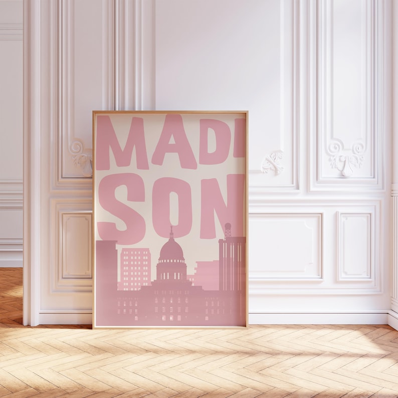 May include: A framed art print featuring the city name "MADISON" in large pink letters above a pink silhouette of the city skyline. The print is displayed in a room with white walls and a wooden floor.