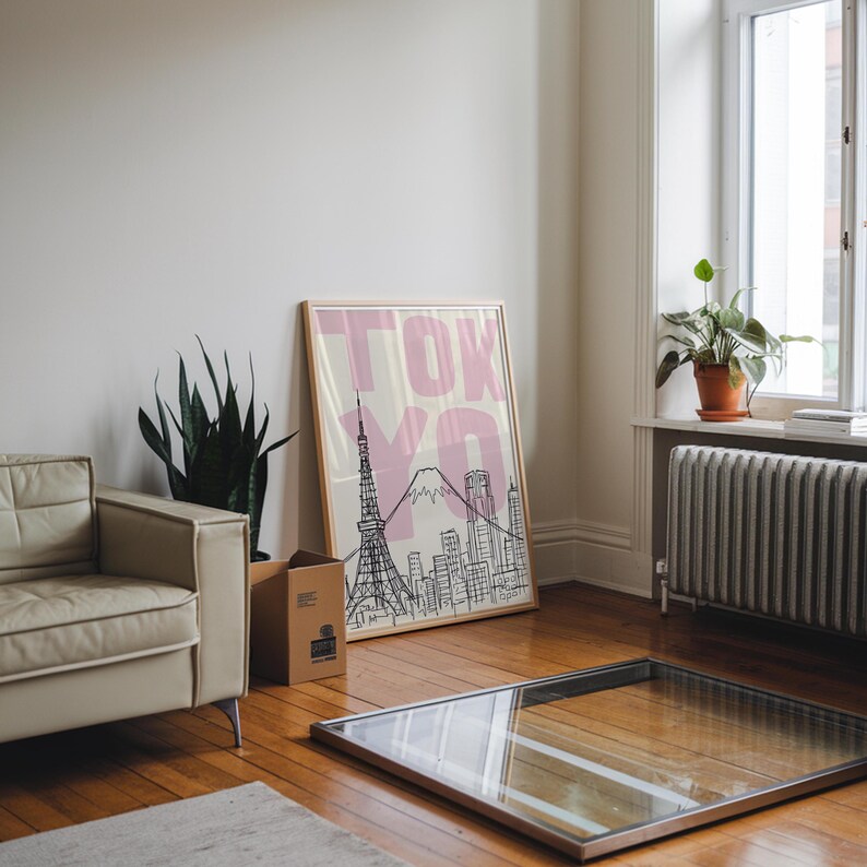 May include: A framed art print featuring a black and white illustration of Tokyo landmarks, including the Tokyo Tower and Mount Fuji, with the word "TOKYO" in pink. The print is displayed in a room with a beige sofa and wooden floor.