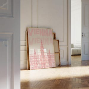 May include: A framed print featuring a pink illustration of a building with a tall clock tower, labeled "VIENNA" in large pink letters. The print is leaning against a wall in a room with a wooden floor.