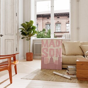 May include: A framed poster with the word "MADISON" in pink block letters, with a pink silhouette of a city skyline and a building. The poster is leaning against a wall in a room with a window, a couch, and a wooden chair.