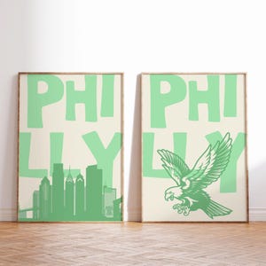 Può includere: Due stampe artistiche incorniciate con una combinazione di colori verde chiaro e crema. Una stampa presenta una silhouette dello skyline di Filadelfia, l'altra mostra un'aquila verde. Entrambe le stampe hanno la parola "PHILLY" in grandi lettere verde chiaro.