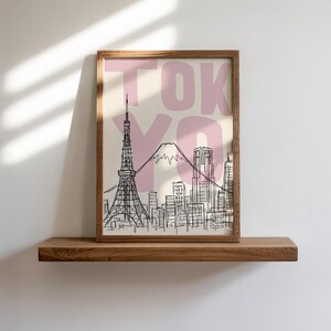 May include: A framed art print featuring a black and white sketch of the Tokyo skyline, including the Tokyo Tower and Mount Fuji. The word "TOKYO" is printed in pink over the image. The print is displayed on a wooden shelf.
