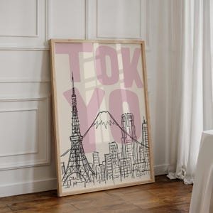 May include: A framed art print featuring a black and white line drawing of Tokyo's skyline, including the Tokyo Tower and Mount Fuji. The word "TOKYO" is printed in large, pink letters over the image. The print is in a light wood frame.