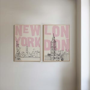 May include: Two framed art prints featuring cityscapes. One print displays the New York City skyline with the Statue of Liberty, and the other showcases London's Big Ben. The words "NEW YORK" and "LONDON" are in pink block letters.