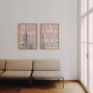 May include: Two framed art prints featuring cityscapes. One print displays the London skyline with the text "LONDON" in pink, and the other showcases the New York skyline with the text "NEW YORK" in pink. The frames are a light brown color.