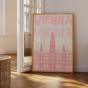 May include: A framed art print featuring the word "VIENNA" in pink, repeated vertically. Below the text is a pink line drawing of a building with a tall central tower and multiple smaller towers. The print is in a wooden frame.