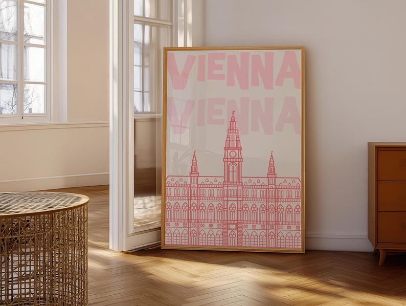 May include: A framed art print featuring the word "VIENNA" in pink, repeated vertically. Below the text is a pink line drawing of a building with a tall central tower and multiple smaller towers. The print is in a wooden frame.