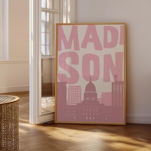 May include: A framed art print featuring the word "MADISON" in large pink letters over a pink and beige cityscape. The artwork includes a silhouette of a capitol building and other buildings.