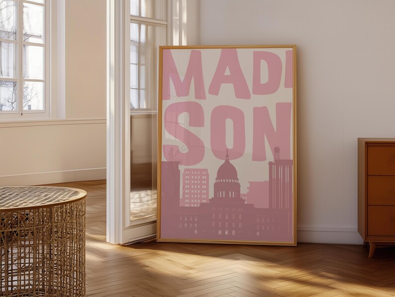 May include: A framed art print featuring the word "MADISON" in large pink letters over a pink and beige cityscape. The artwork includes a silhouette of a capitol building and other buildings.