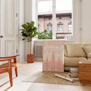 May include: A framed art print featuring a pink illustration of a building with the word "VIENNA" in pink text. The print is displayed in a living room setting with a beige sofa, wooden furniture, and a large potted plant.