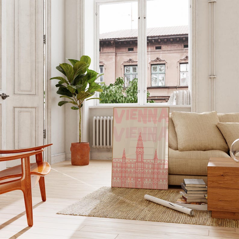 May include: A framed art print featuring a pink illustration of a building with the word "VIENNA" in pink text. The print is displayed in a living room setting with a beige sofa, wooden furniture, and a large potted plant.