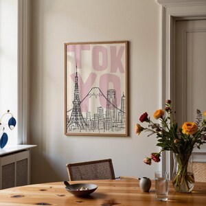 May include: A framed art print featuring a black and white line drawing of the Tokyo skyline, including the Tokyo Tower and Mount Fuji. The word "TOKYO" is printed in pink block letters above the illustration. The print is displayed in a room with a wooden table and a vase of flowers.
