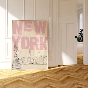 May include: A framed art print featuring the word "NEW YORK" in pink block letters above a black and white skyline illustration. The Statue of Liberty is visible on the left. The print is displayed in a room with a wooden floor.