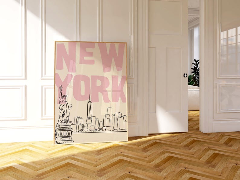 May include: A framed art print featuring the word "NEW YORK" in pink block letters above a black and white skyline illustration. The Statue of Liberty is visible on the left. The print is displayed in a room with a wooden floor.
