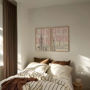 May include: Two framed prints with the words "LONDON" and "NEW YORK" in pink, above line drawings of Big Ben and the Statue of Liberty. The prints hang above a bed with a striped comforter, white pillows, and brown throw pillows.