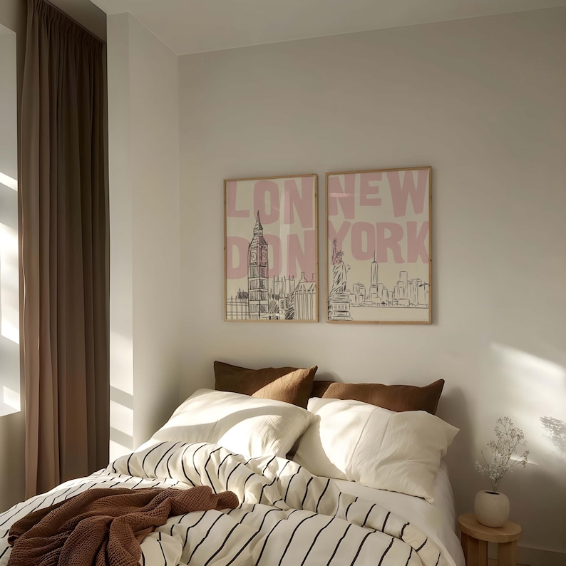 May include: Two framed prints with the words "LONDON" and "NEW YORK" in pink, above line drawings of Big Ben and the Statue of Liberty. The prints hang above a bed with a striped comforter, white pillows, and brown throw pillows.
