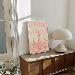 May include: A framed art print featuring the word "VIENNA" in pink, above a pink line drawing of a building. The print is displayed on a wooden cabinet, next to a white lamp with a rounded shade.