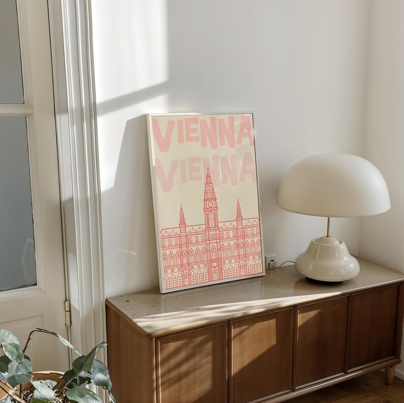 May include: A framed art print featuring the word "VIENNA" in pink, above a pink line drawing of a building. The print is displayed on a wooden cabinet, next to a white lamp with a rounded shade.