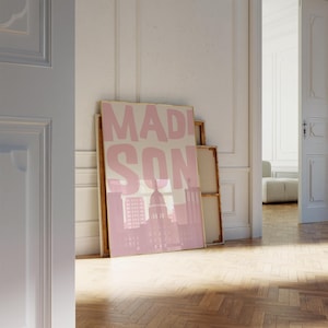 May include: A framed pink and beige art print featuring the word "MADISON" in large block letters, with a pink silhouette of a city skyline. The artwork is leaning against a wall in a room with a wooden floor.