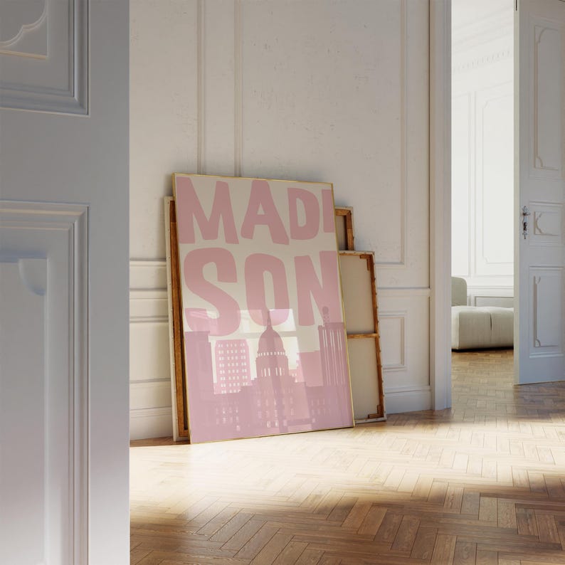 May include: A framed pink and beige art print featuring the word "MADISON" in large block letters, with a pink silhouette of a city skyline. The artwork is leaning against a wall in a room with a wooden floor.