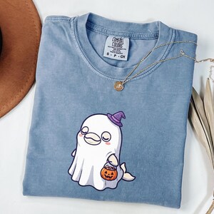 May include: A blue short-sleeved t-shirt featuring a graphic of a white ghost character wearing a purple witch hat and holding an orange jack-o'-lantern candy bucket. The shirt has a crew neckline and a Comfort Colors brand label inside.
