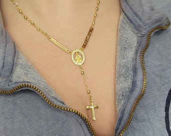 May include: A gold-toned necklace with a small oval pendant featuring a religious figure. The pendant is surrounded by small clear stones. The necklace also includes two small rectangular charms with the word "Siempre" engraved on them. The necklace is worn by a person wearing a blue shirt.