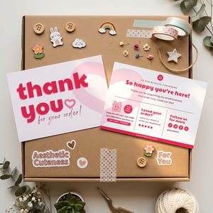 May include: A brown cardboard shipping box decorated with stickers, buttons, and washi tape. Two white cards are placed on top, one reading thank you for your order and the other reading So happy you're here with social media icons and text.