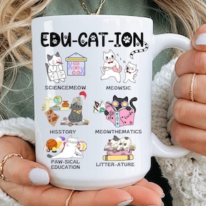 Puede incluir: Taza de cerámica blanca con el texto "EDU-CAT-ION" e ilustraciones de gatos que representan diferentes materias escolares. La taza es sostenida por una persona que lleva un suéter blanco.