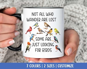 May include: White ceramic mug with a black rim and handle. The mug features a colorful illustration of birds and the text "Not all who wander are lost, some are just looking for birds".