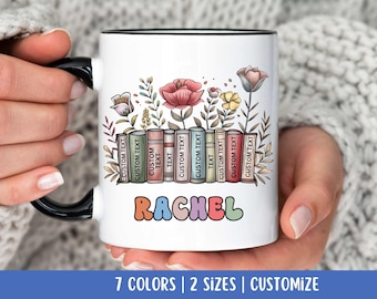 Puede incluir: Taza de cerámica blanca con asa negra. La taza presenta una ilustración colorida de una pila de libros con flores y el nombre "RACHEL" en letras de colores.