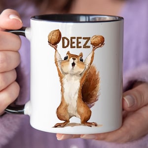 Puede incluir: Taza de cerámica blanca con interior y asa negros. La taza presenta una ardilla de dibujos animados sosteniendo dos nueces sobre su cabeza, con la palabra "DEEZ" impresa sobre la ardilla en letras marrones.