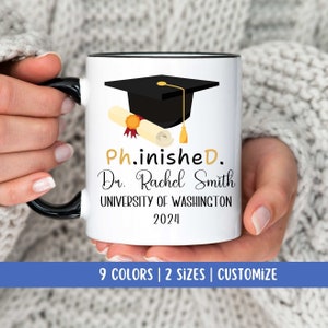 May include: White coffee mug with a black rim and handle. The mug features a black graduation cap with a gold tassel and a rolled up diploma with a gold seal. The text on the mug reads "Ph.inisheD. Dr. Rachel Smith UNIVERSITY OF WASHINGTON 2024".
