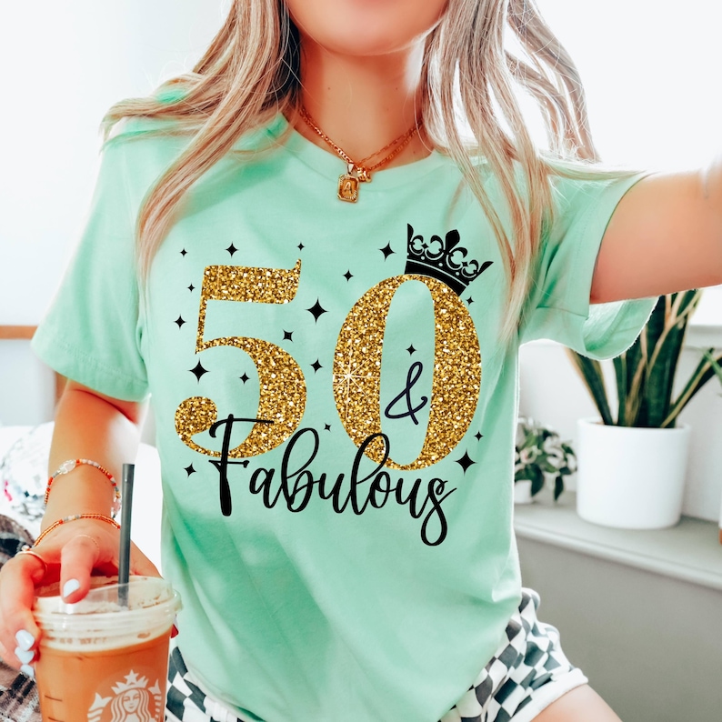 May include: A mint green short-sleeve t-shirt featuring a graphic design with the text 50 & Fabulous in gold glitter font. The design includes a black crown icon above the zero, surrounded by small black star accents on the front of the shirt.