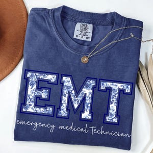 Puede incluir: Una camiseta azul marino con las letras "EMT" en una fuente en negrita, delineada y rellena con un estampado floral azul. Las palabras "emergency medical technician" est&aacute;n escritas debajo en una fuente cursiva. Un collar dorado con un colgante est&aacute; sobre la camiseta.