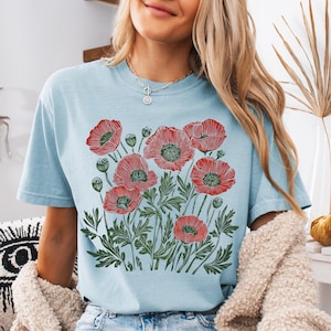 May include: Light blue t-shirt featuring a red and green poppy flower design. The graphic includes detailed illustrations of poppy blooms, buds, and leaves. The shirt is a short-sleeved, crew neck style.