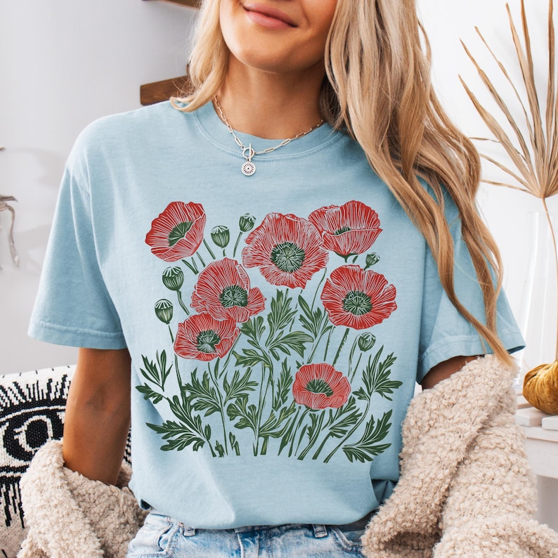 May include: Light blue t-shirt featuring a red and green poppy flower design. The graphic includes detailed illustrations of poppy blooms, buds, and leaves. The shirt is a short-sleeved, crew neck style.