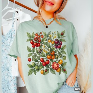 Op de afbeelding: Lichtgroen T-shirt met een aquarel illustratie van een tomatenplant. Het ontwerp bevat rode, oranje en groene tomaten, groene bladeren, gele bloemen, een bij en lieveheersbeestjes. Het woord "BAY" staat rechtsonder.