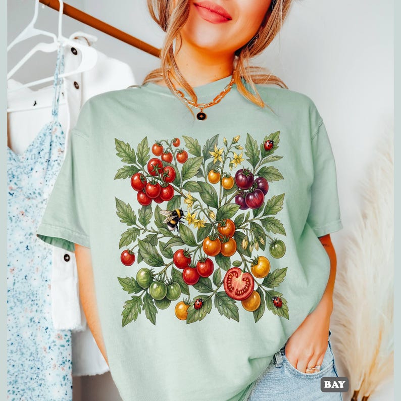 Op de afbeelding: Lichtgroen T-shirt met een aquarel illustratie van een tomatenplant. Het ontwerp bevat rode, oranje en groene tomaten, groene bladeren, gele bloemen, een bij en lieveheersbeestjes. Het woord "BAY" staat rechtsonder.