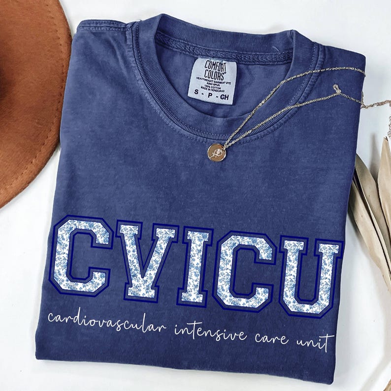 May include: A navy blue t-shirt with the letters "CVICU" in a floral pattern, with the words "cardiovascular intensive care unit" below. A gold necklace with a pendant rests on the shirt.