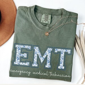 Puede incluir: Camiseta verde oliva con las letras "EMT" en un dise&ntilde;o floral azul. Las palabras "emergency medical technician" est&aacute;n impresas debajo. Un collar dorado con un colgante descansa sobre la camiseta doblada.