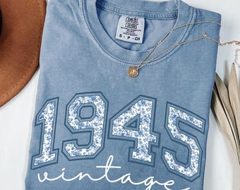 Può includere: Una maglietta blu Comfort Colors con l'anno "1945" in un motivo floreale e la parola "vintage" in corsivo bianco. Una collana d'oro con un ciondolo è drappeggiata sulla maglietta.