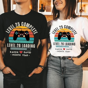 May include: A couple's matching t-shirts with a retro video game controller graphic. The shirts say "Level 25 Complete" and "Level 26 Loading" with a progress bar. The shirts are personalized with the names "Kaken" and "David" and the month and year.