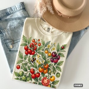Op de afbeelding: Ivoor kleurig T-shirt met een ontwerp van een tomatenplant met rode, oranje, gele en groene tomaten, samen met een bij en een lieveheersbeestje. Het shirt wordt gecombineerd met een blauwe jeans en een beige hoed.