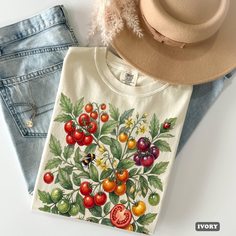 Op de afbeelding: Ivoor kleurig T-shirt met een ontwerp van een tomatenplant met rode, oranje, gele en groene tomaten, samen met een bij en een lieveheersbeestje. Het shirt wordt gecombineerd met een blauwe jeans en een beige hoed.
