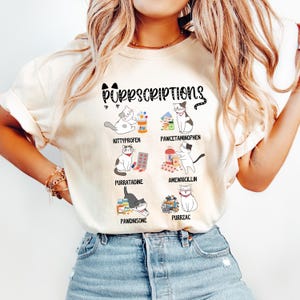 Könnte beinhalten: Cremefarbenes T-Shirt mit dem Text "Purrscriptions" und Illustrationen von Katzen mit Medikamenten. Das Shirt zeigt die Namen der Medikamente: Kittyprofen, Pawcetaaminophen, Purratadine, Amewxicillin, Pawdnisone und Purrzac.